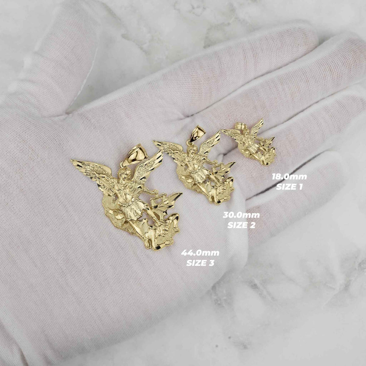 Three yellow gold Saint Michael pendants of varying sizes held in a gloved hand with labels.