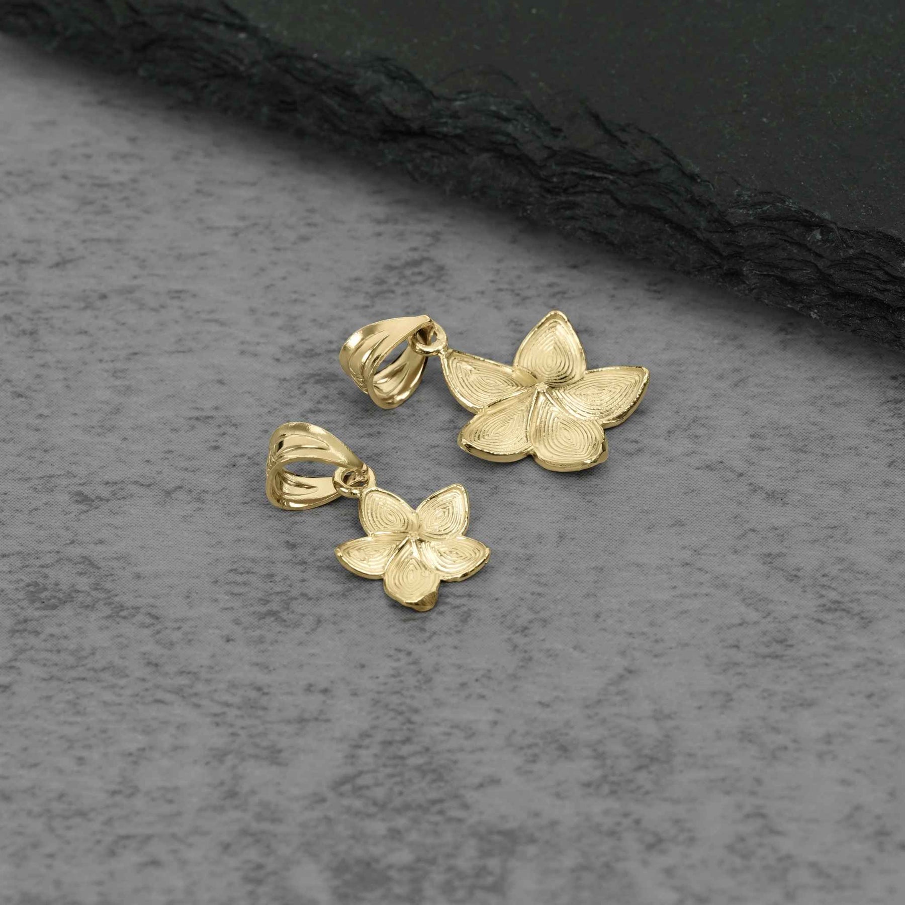 Two gold Fiori hibiscus flower pendant charms displayed on a textured slate surface.