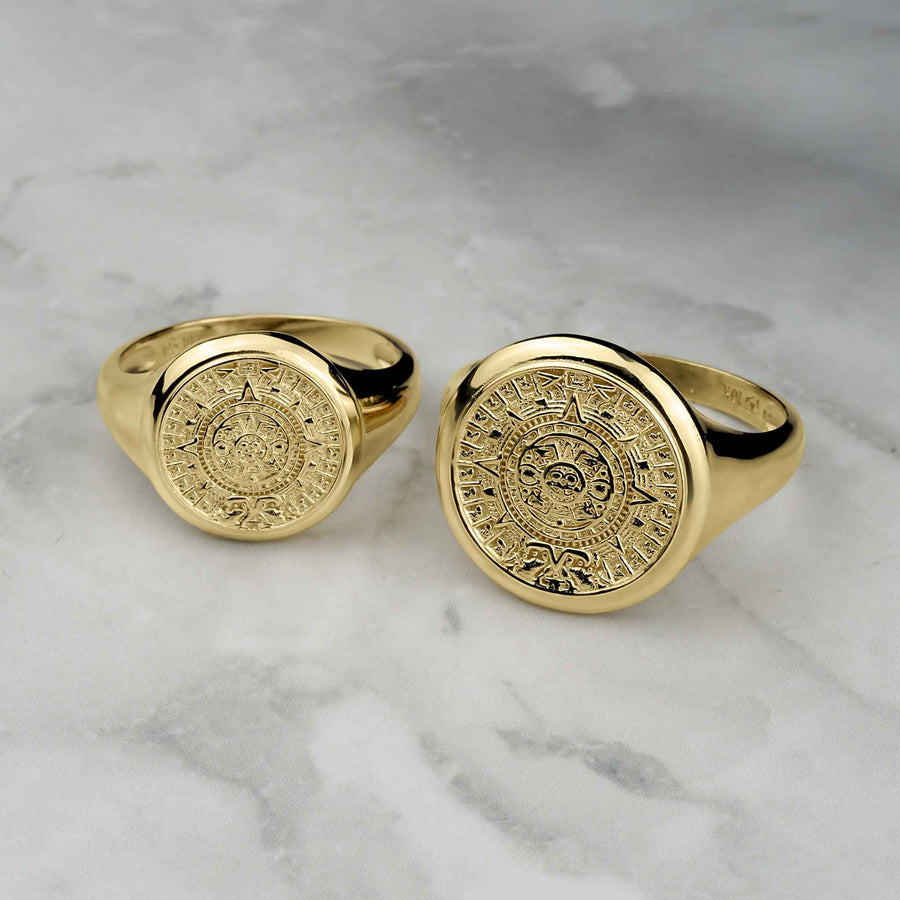 Two round yellow gold signet rings featuring detailed Aztec Mayan calendar designs on marble.