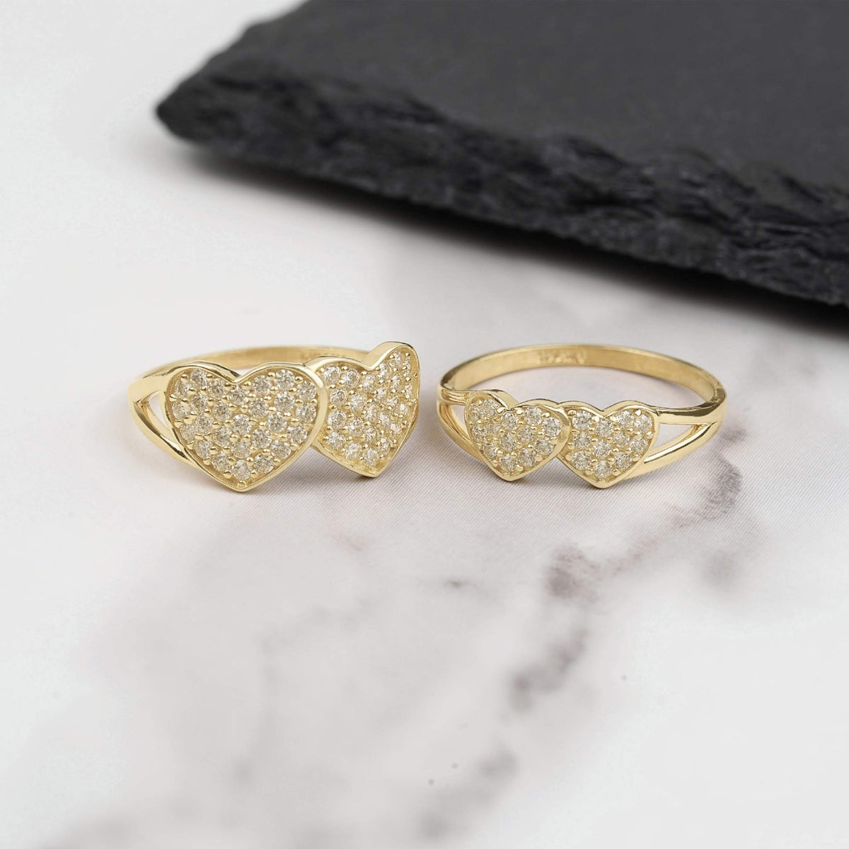 Two gold dual heart rings with pavé white CZ stones on white marble surface