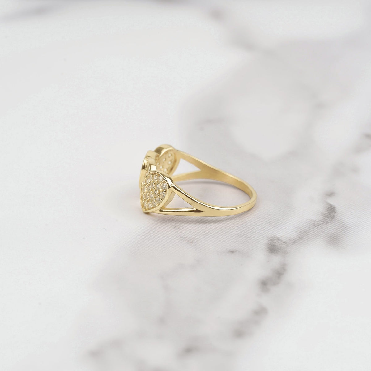 Side profile view of gold dual heart ring with pavé white CZ stones on white marble surface
