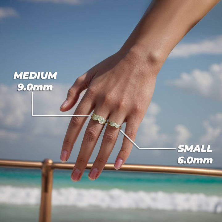 On-hand view of two pave-set gold heart rings of different sizes against a beach backdrop.
