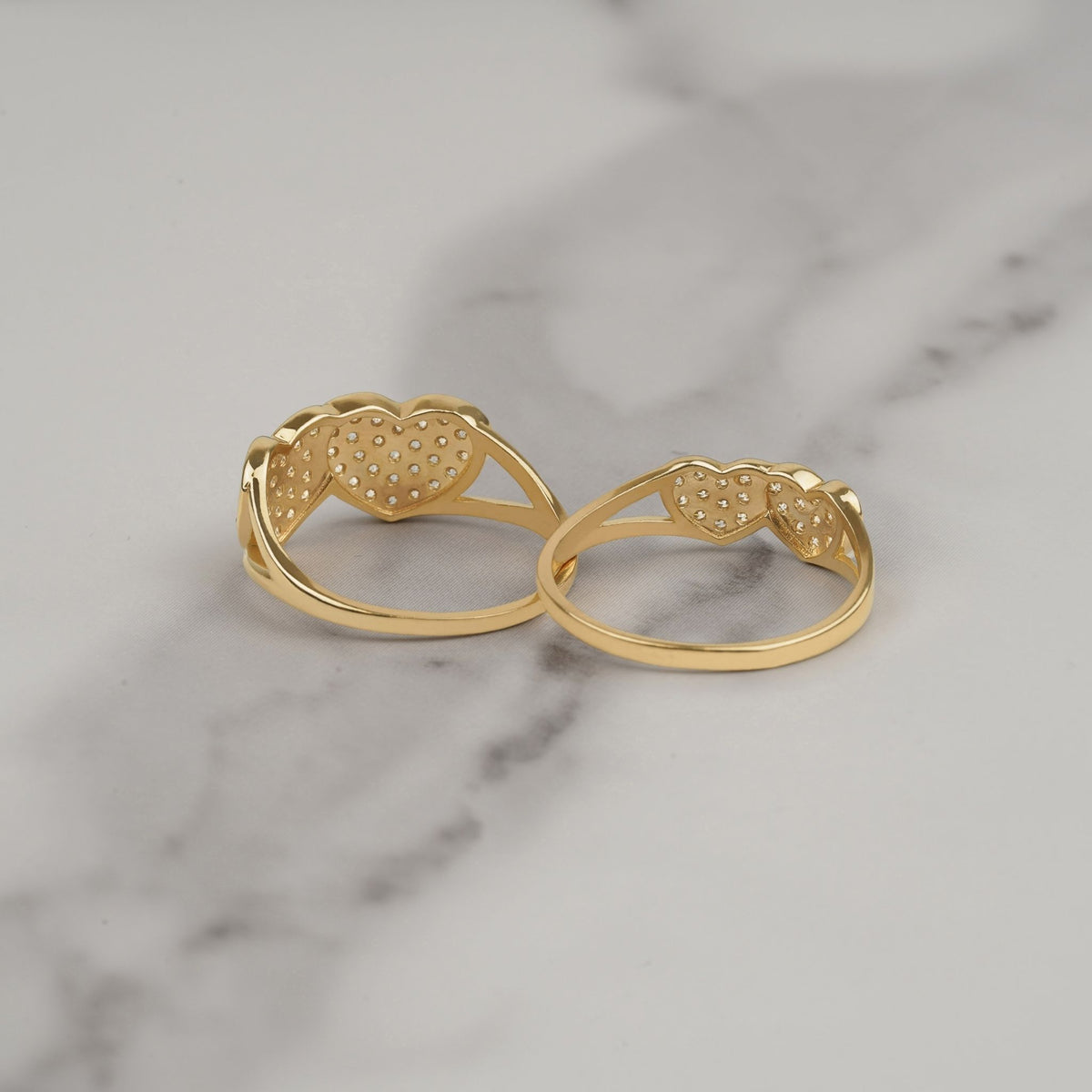 Back view of two gold dual-heart cubic zirconia rings on white marble.