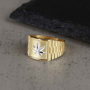 Gold Rolex-style ring with a silver cannabis leaf design on a gray textured surface.