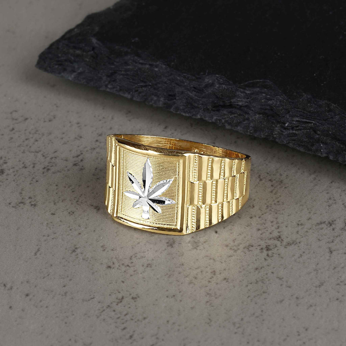 Gold Rolex-style ring with a silver cannabis leaf design on a gray textured surface.