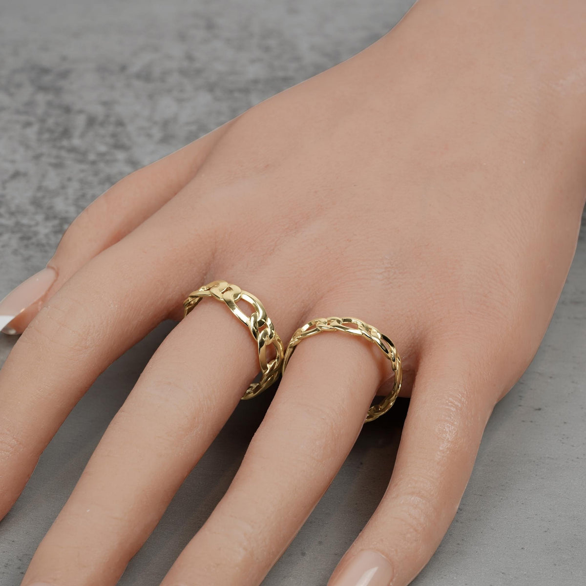 Hand wearing two solid gold Figaro chain-link rings on a neutral surface.