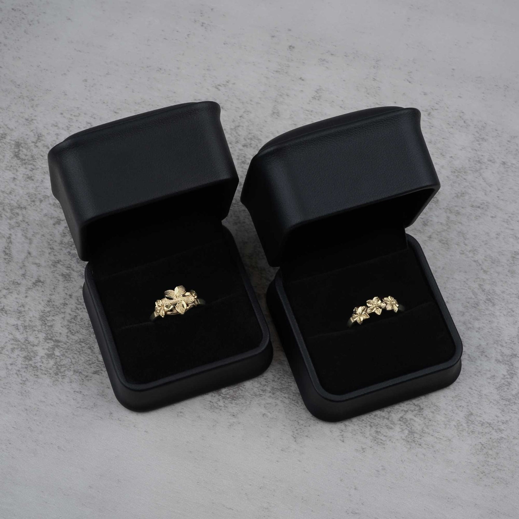 Two black jewelry boxes with 10k- 14k gold rings with floral design on a gray surface