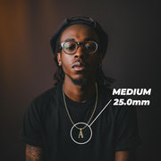 Model wearing a medium 25mm gold initial 'A' pendant labeled MEDIUM 25.0mm on a black shirt.