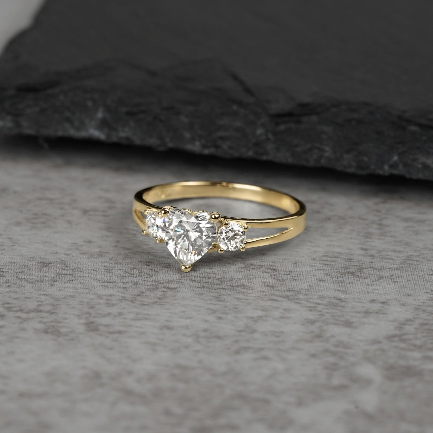 Three-stone gold ring with heart-shaped white cubic zirconia on gray background.