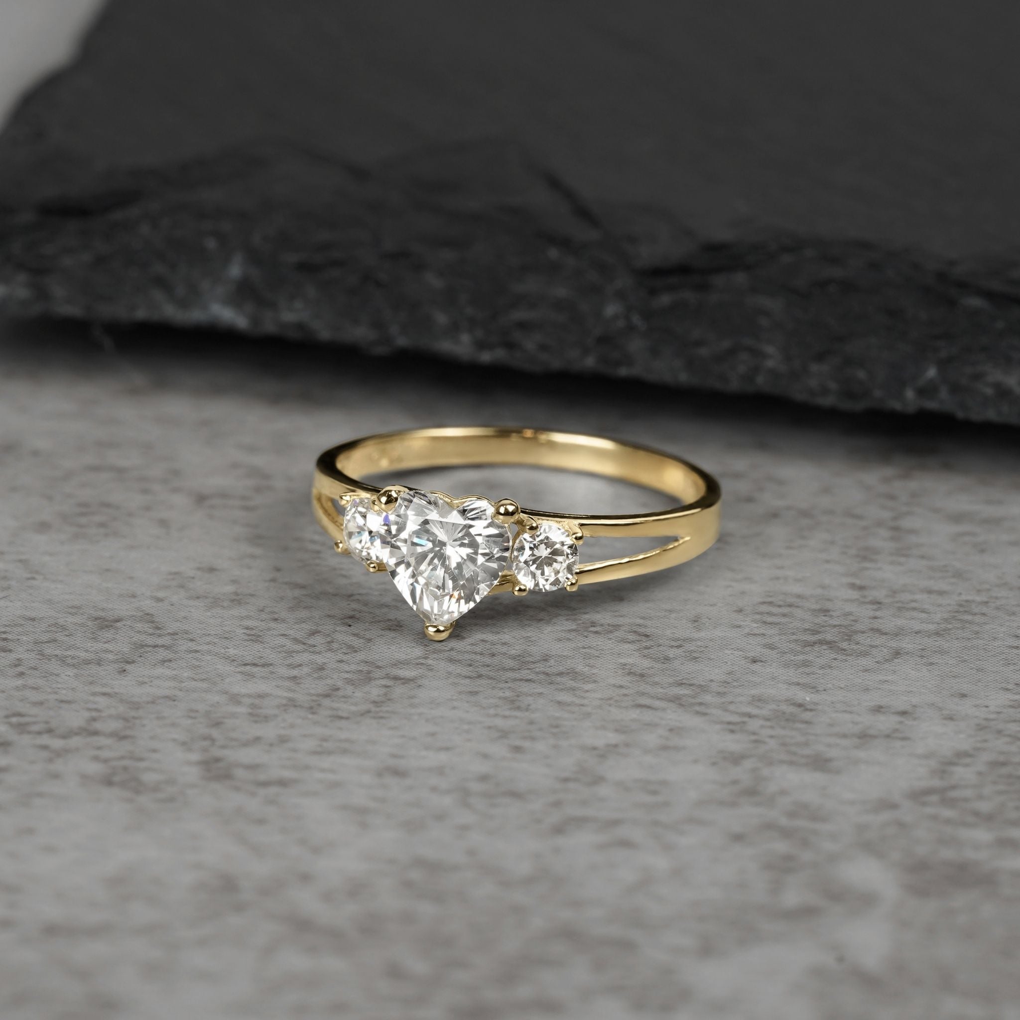 Three-stone gold ring with heart-shaped white cubic zirconia on gray background.