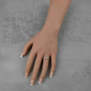 A mannequin hand with manicured nails wearing a gold infinity ring against gray concrete.