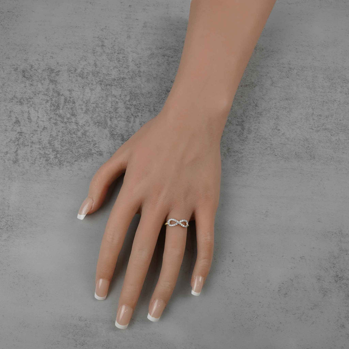 A mannequin hand with manicured nails wearing a gold infinity ring against gray concrete.