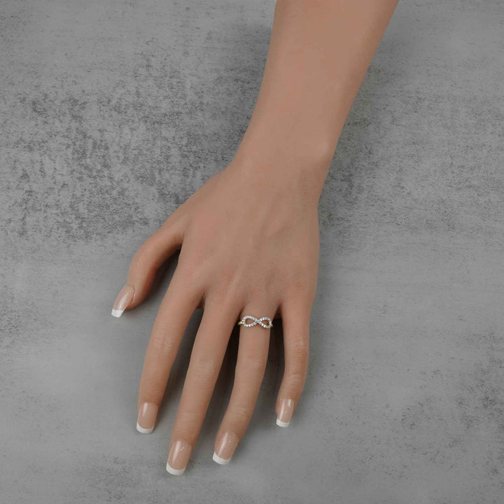 A mannequin hand with manicured nails wearing a gold infinity ring against gray concrete.