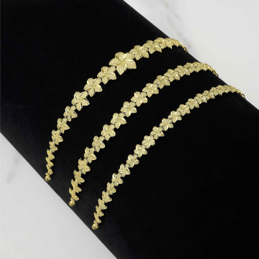 Three gold hibiscus flower bracelets in varying widths displayed on a black surface.