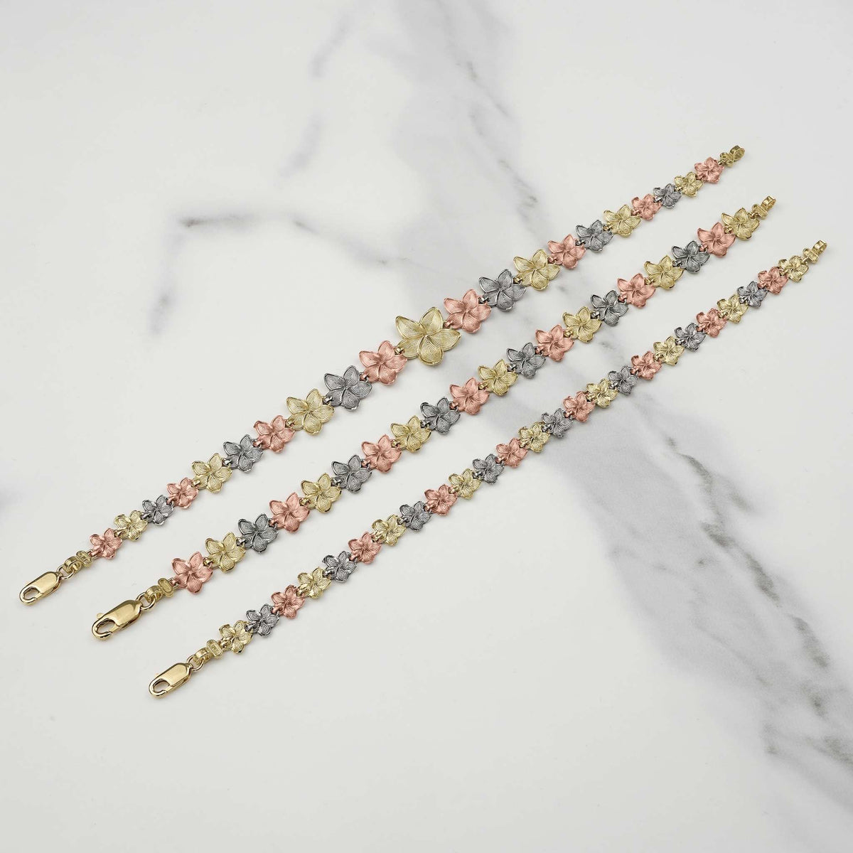 Three gold Fiori hibiscus flower link bracelets laid diagonally on white marble