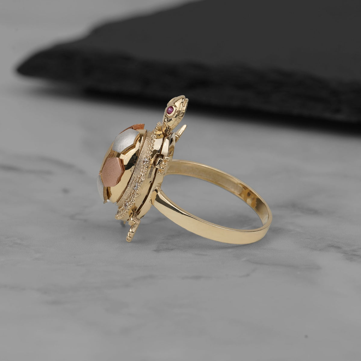 Side view of a tri-color gold turtle locket ring on a marble surface.