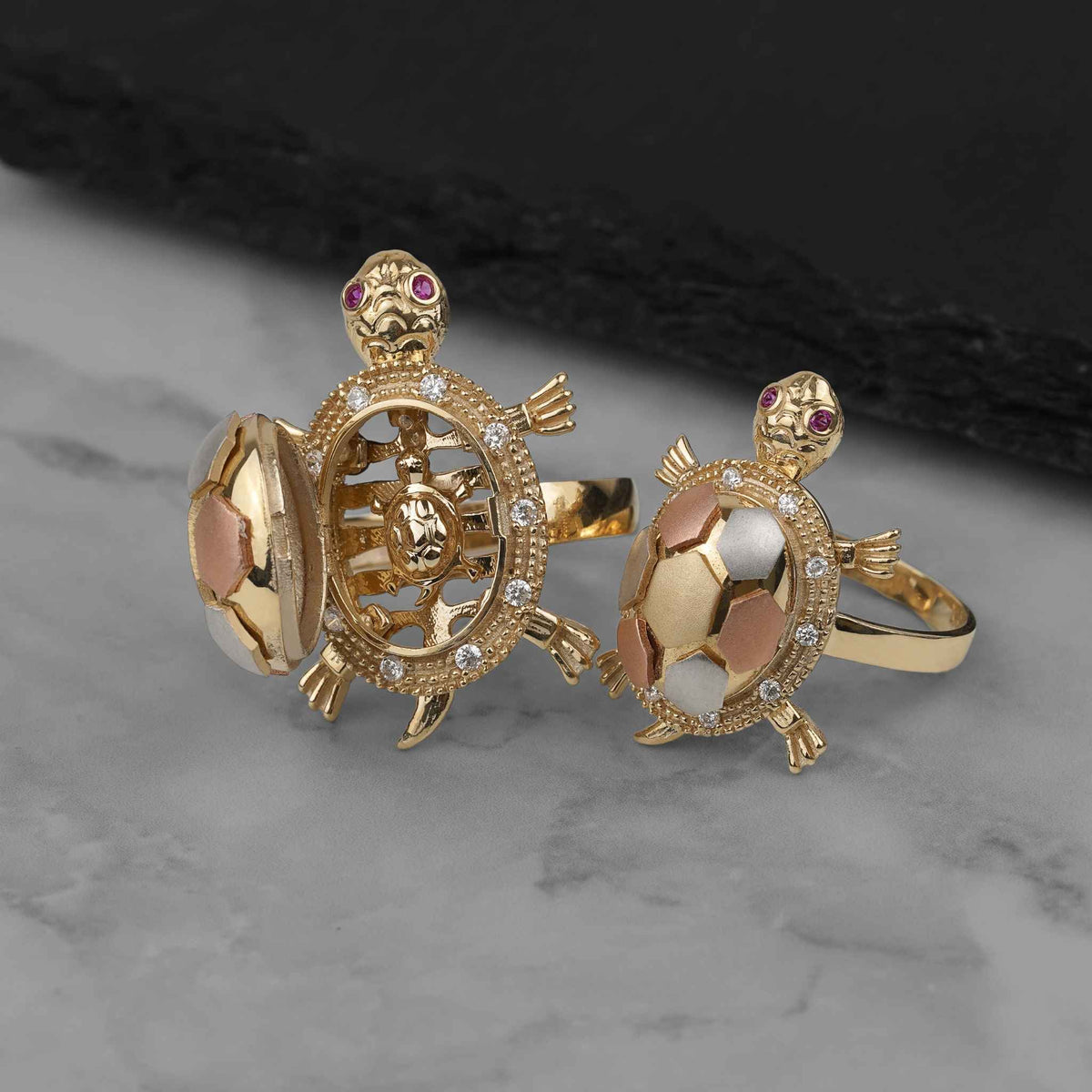 Two gold turtle locket rings on a marble surface, one open to reveal detailed interioring.