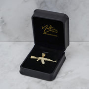 A gold-tone AK-47 pendant displayed inside an open black jewelry box on a marble surface.