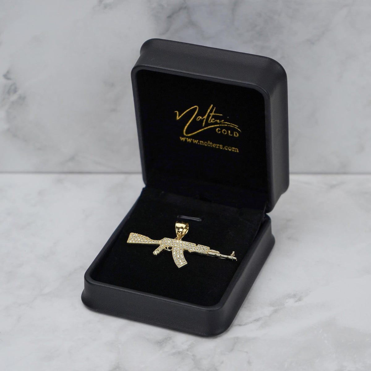 A gold-tone AK-47 pendant displayed inside an open black jewelry box on a marble surface.