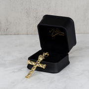 Gold cross pendant resting inside an open black jewelry box with gold branding.