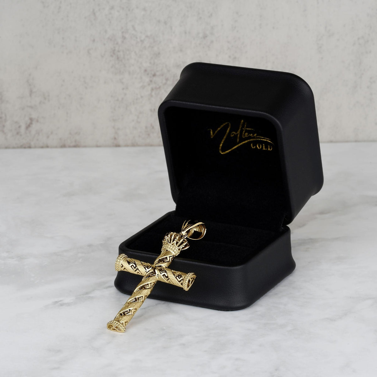 Gold cross pendant resting inside an open black jewelry box with gold branding.