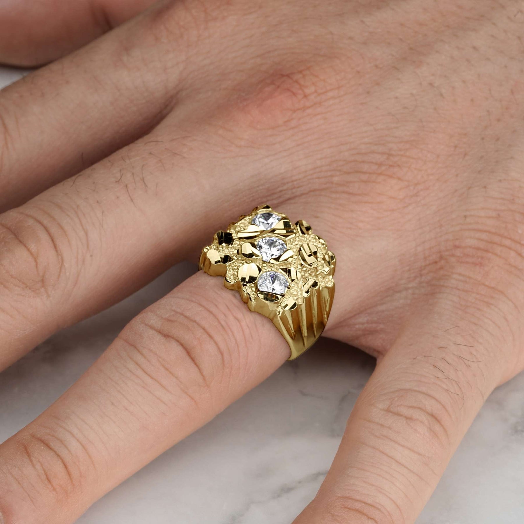 A heavy, diamond-cut yellow gold nugget ring with three shimmering gemstones worn on a finger.