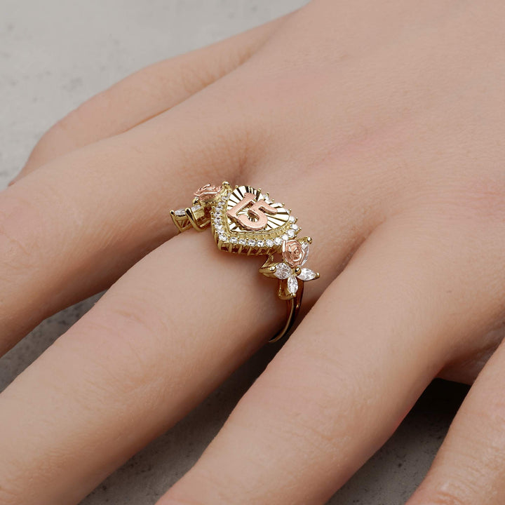 A gold two-tone Quinceañera 15 heart ring with rose details being worn on a finger.
