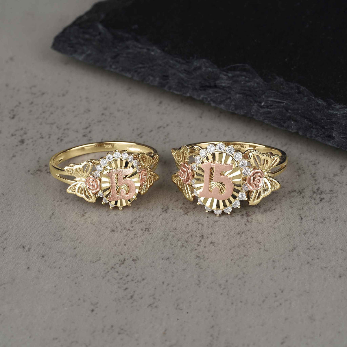 Two sizes of the gold two-tone Quinceañera 15 ring displayed on a gray surface.