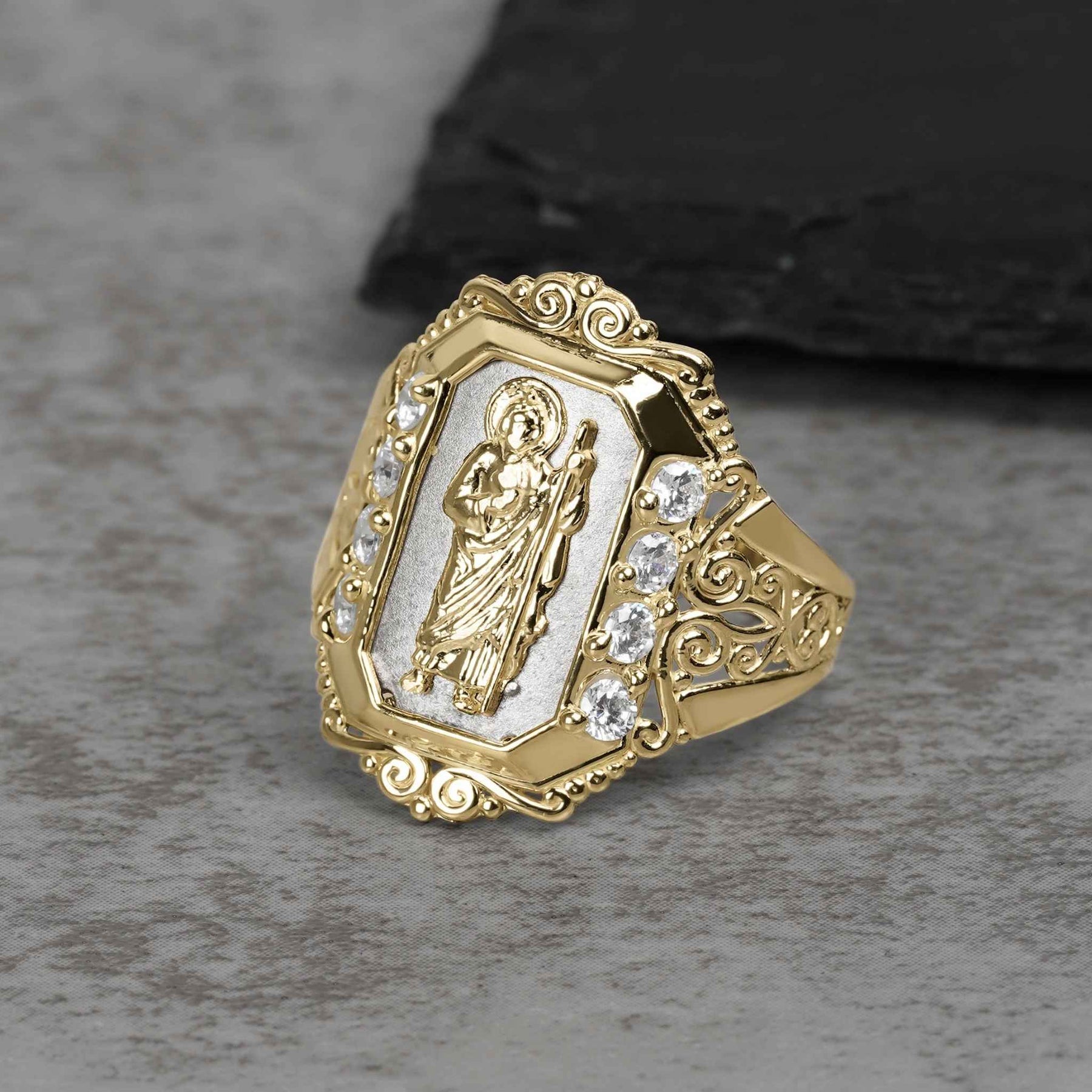 Two-tone gold San Judas royal ring featuring cubic zirconia stones on a gray background.