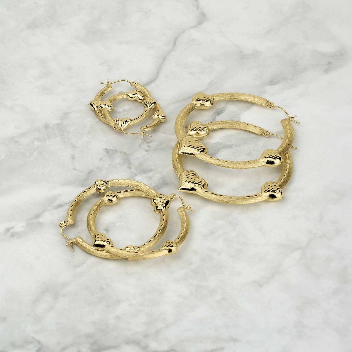 Three different sizes of textured gold tre heart hoop earrings on a marble surface.