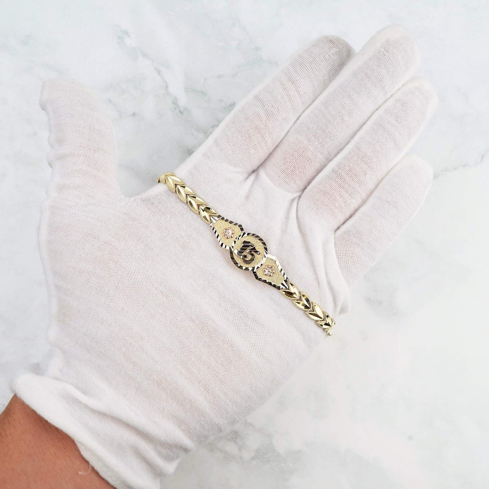 Tri-color gold quinceañera bracelet featuring a central "15" medallion, displayed on a white-gloved hand.