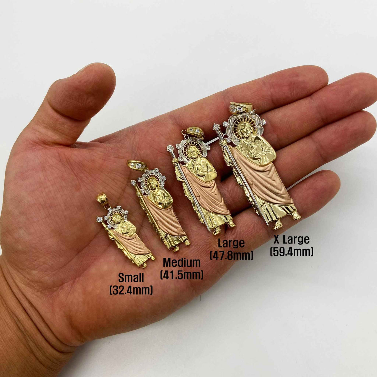 Hand holding four sizes of tri-color gold Saint Jude Thaddeus religious pendants with size labels.