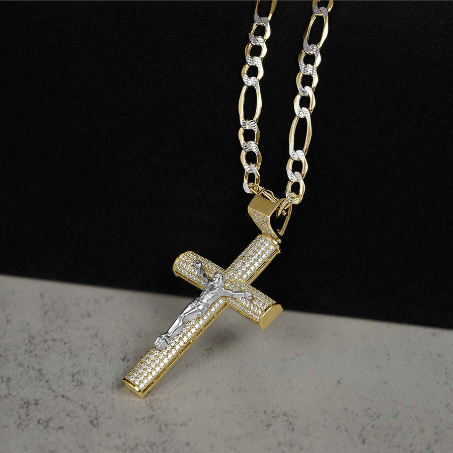 Angled view of two-tone gold Hermo iced crucifix pendant on a pave Figaro chain resting on a dark stone surface.