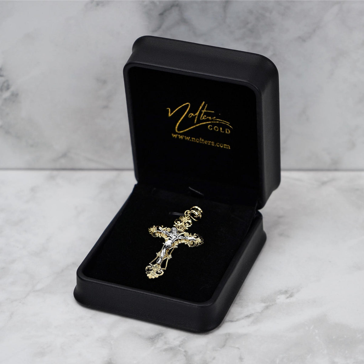 Ornate gold cross pendant displayed in a black velvet-lined jewelry box on a marble surface.