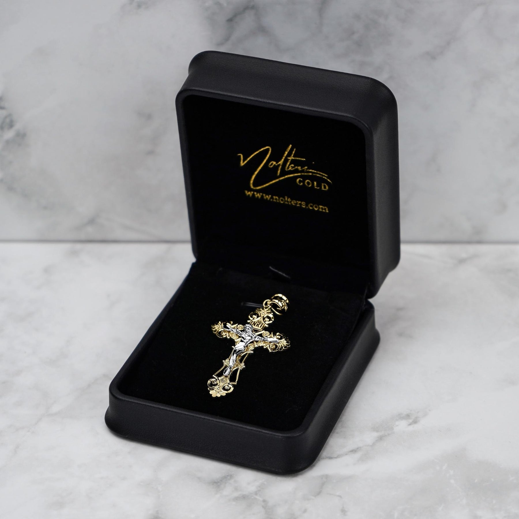Gold crucifix pendant displayed inside an open black jewelry box with gold branding.