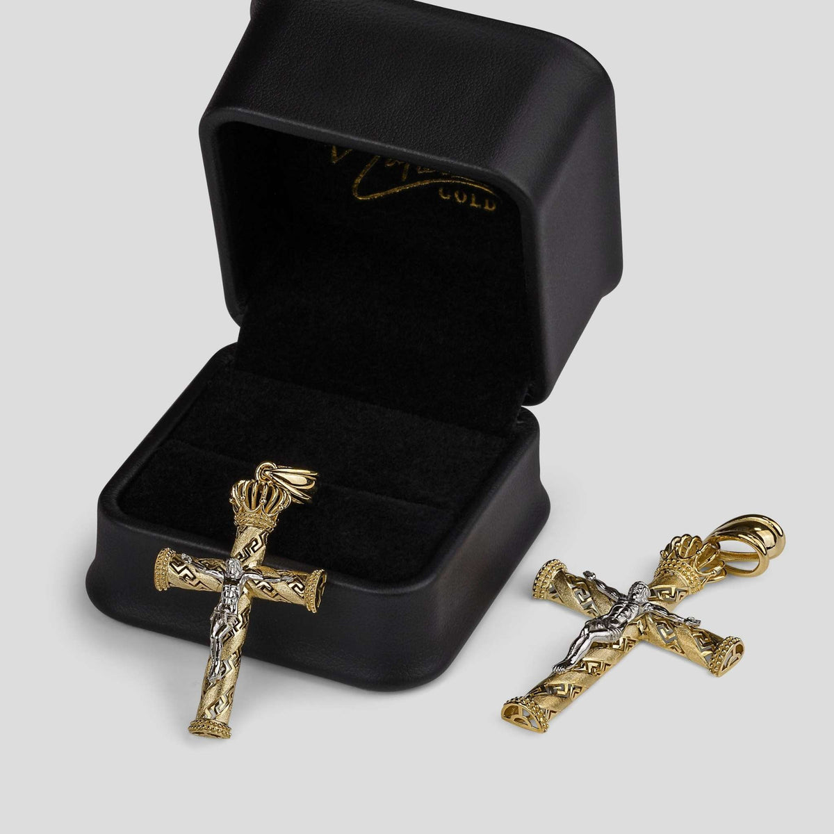 Two gold crowned crucifix pendants displayed next to an open black jewelry box.