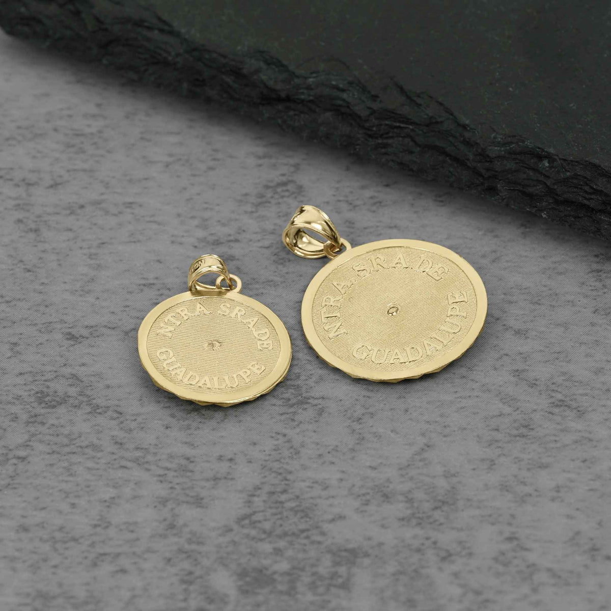 Two gold-toned round pendant charms with bail loops, featuring engraved lettering on a textured gray surface.
