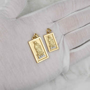 Two rectangular gold Virgin Mary Guadalupe pendants displayed on a white gloved hand.