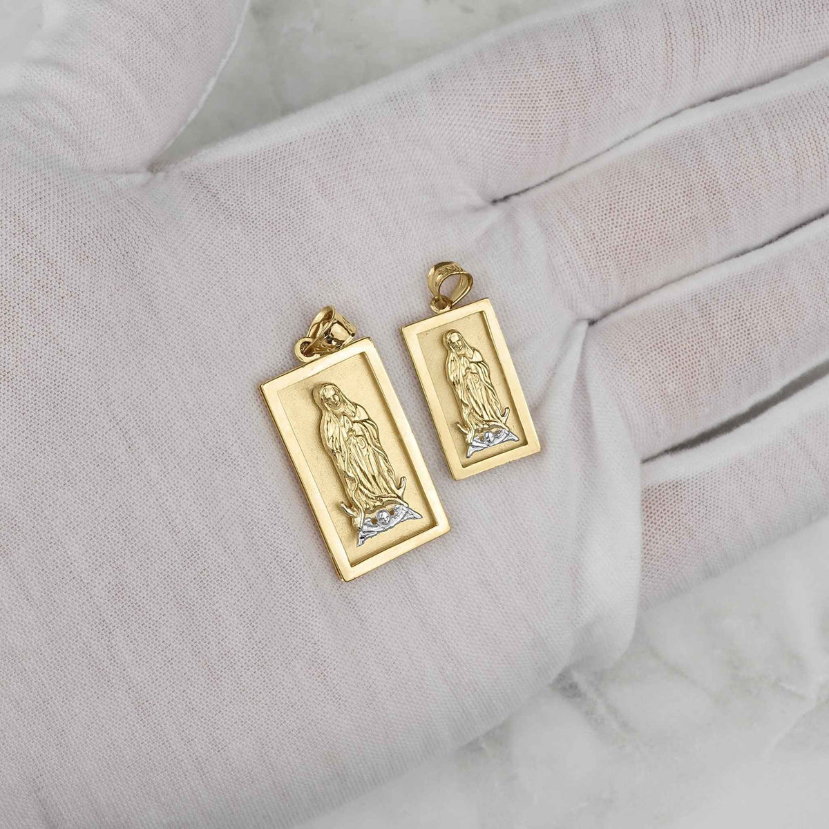Two rectangular gold Virgin Mary Guadalupe pendants displayed on a white gloved hand.