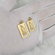 Two rectangular gold Guadalupe pendants resting on a white fabric-covered hand.