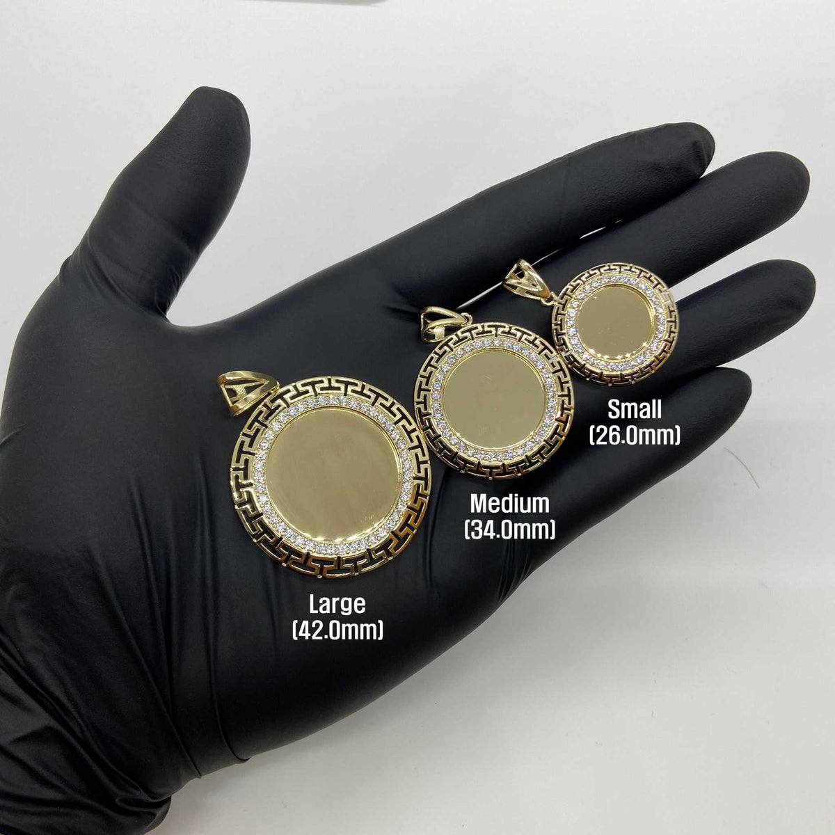 Gloved hand holding three sizes of gold Greek key CZ round photo pendant charms.