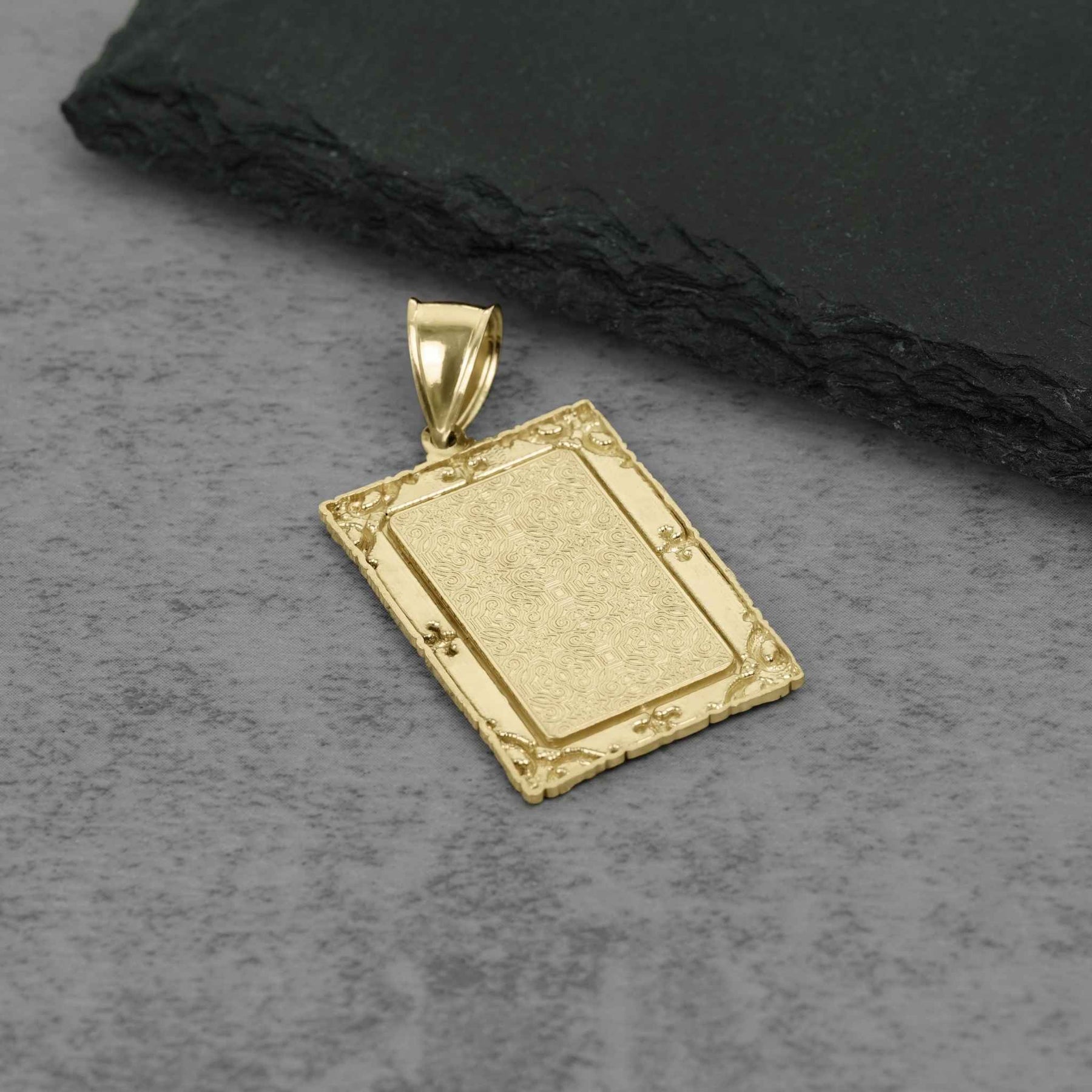Solid gold memorial picture pendant with intricate decorative borders displayed on gray stone.