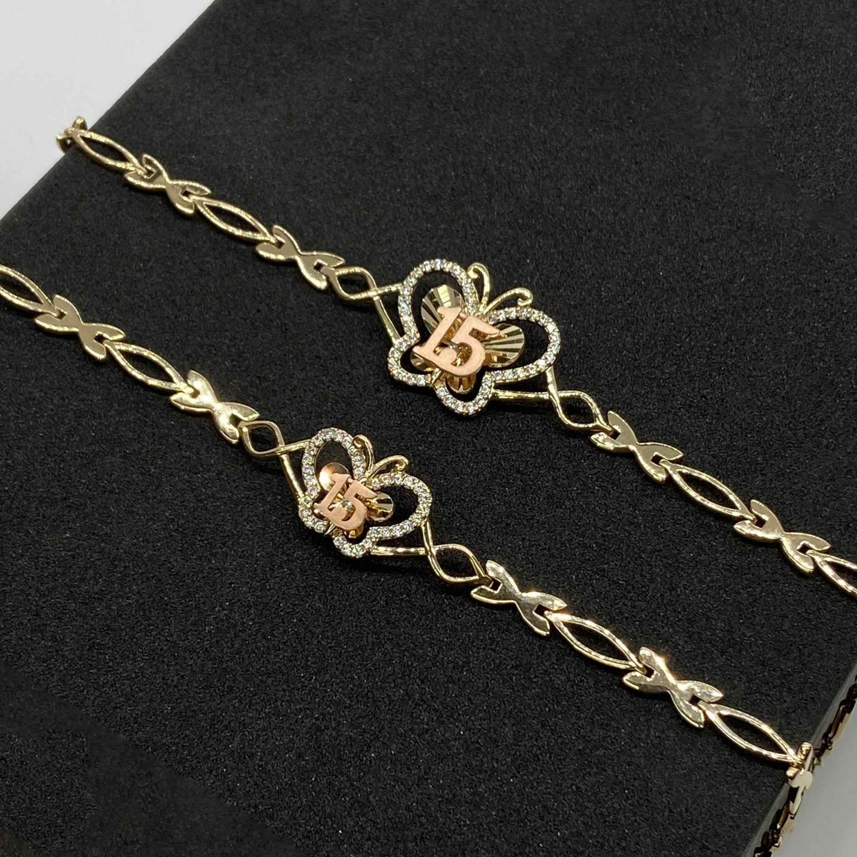 Two gold Quinceañera 15 butterfly charm bracelets with cubic zirconia accents on black display
