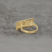 Back view of a gold Last Supper signet ring on a gray textured surface.