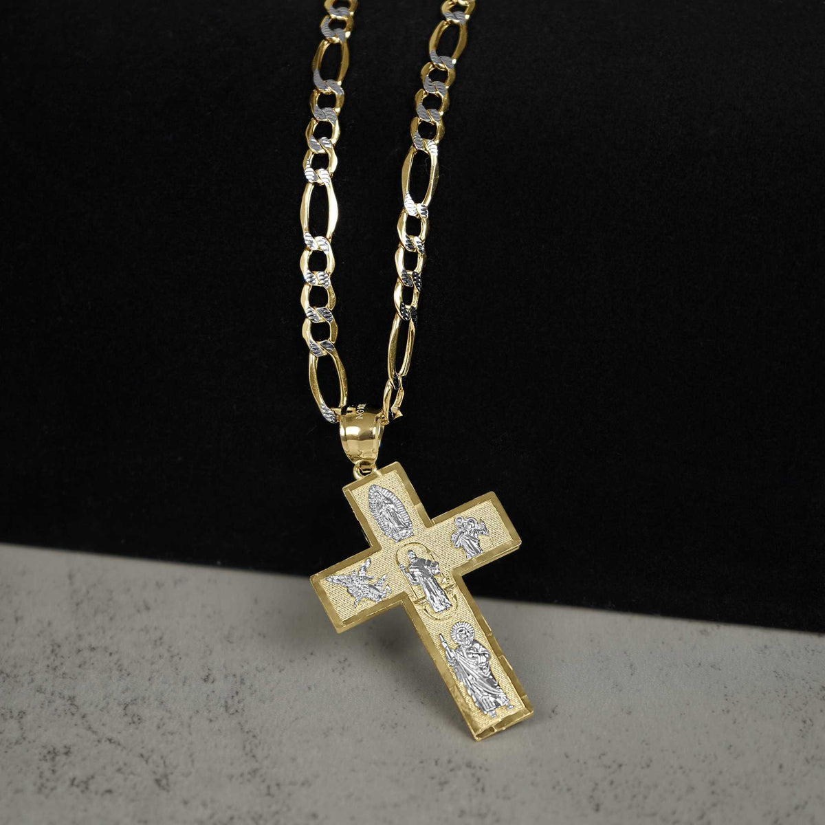 Gold cross pendant with silver-tone religious engravings, hanging from a gold Figaro chain against a two-tone background.
