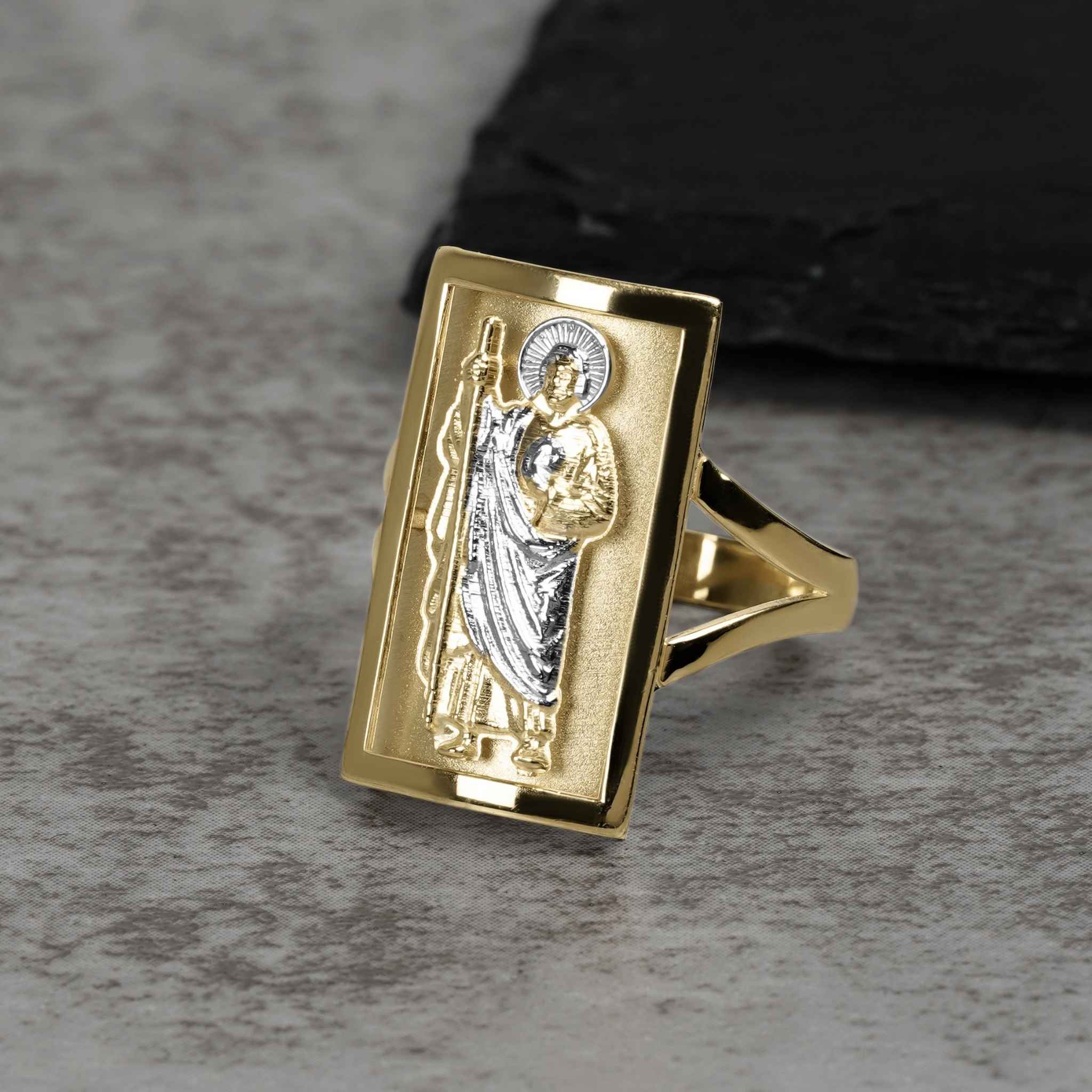 Men's two-tone gold rectangular San Judas signet ring displayed on a textured surface.