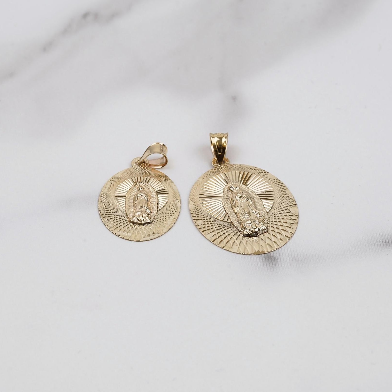 Two gold Virgin Mary oval pendants in different sizes on a marble background.