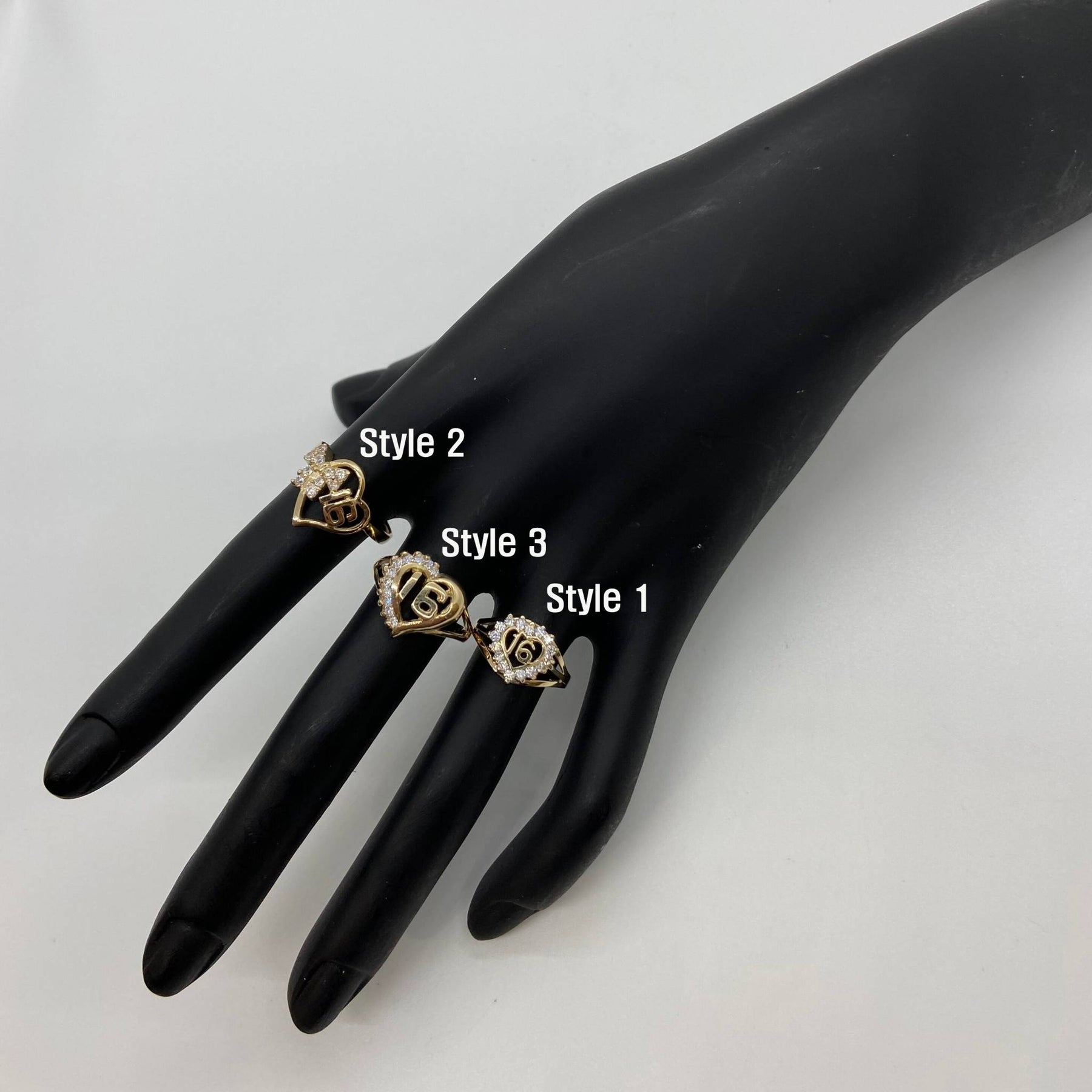 Black mannequin hand wearing three gold Sweet 16 Heart Ring with different designs.