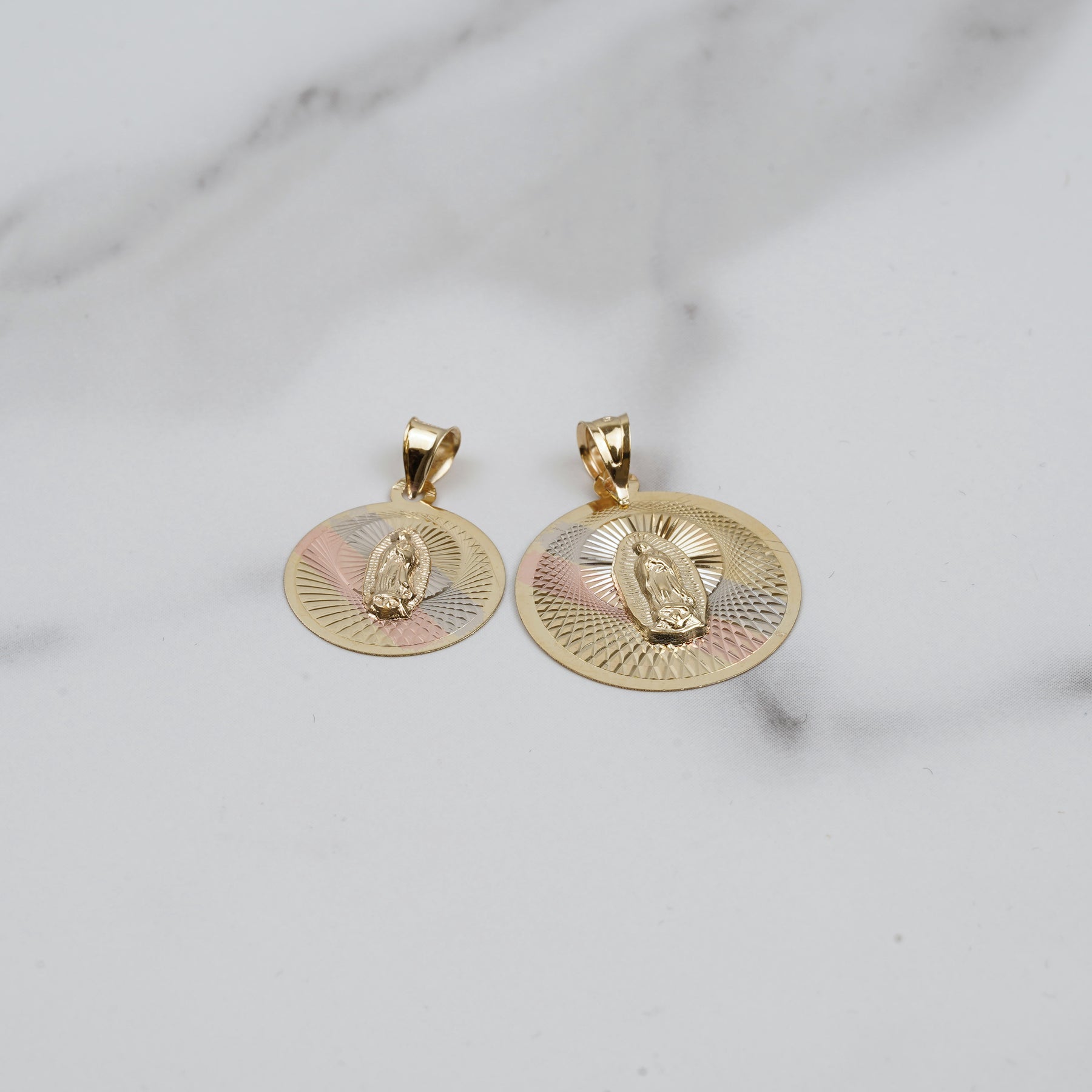 Two tricolor gold round pendants featuring Virgin Mary engravings and sunburst patterns on a white marble surface.