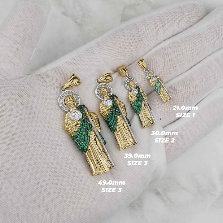 Four gold and green San Judas pendants on white fabric showing different size labels.